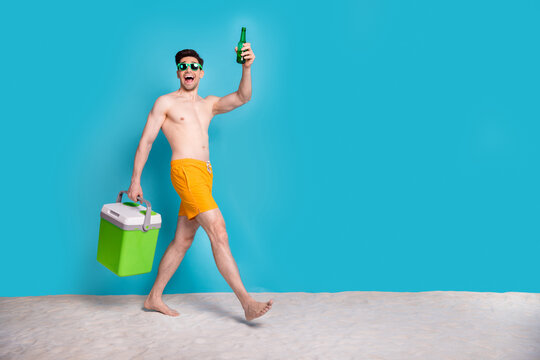 Full size photo of attractive young man walk hold beer bottle carry fridge wear trendy yellow shorts isolated on blue color background