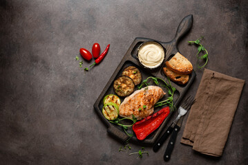 Grilled chicken breast with grilled vegetables, toast and sauce on a wooden cutting board. Dark rustic background. Top view, flat lay, copy space