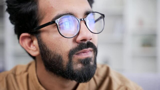 Close up portrait of young handsome bearded businessman in glasses sitting in room at home office looking away. Head shot of serious thinking IT programmer developer Confident entrepreneur in shirt