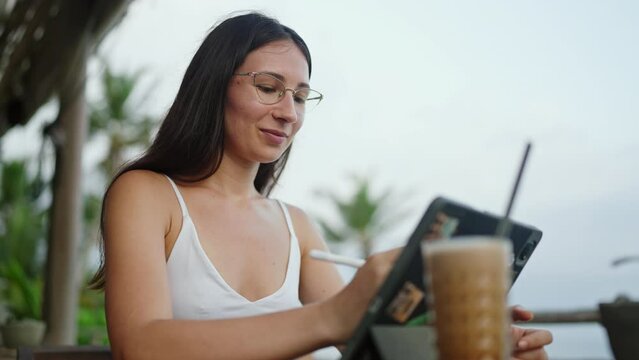 Female freelancer draws with stylus on a graphics tablet in cafe with ocean view, palm trees. Artist girl working on device by the seaside. Young illustrator on remote work day on tropical island.