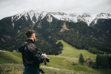 Fotógrafo  con cámara en mano observando paisaje de montañas nevadas y prado verde.