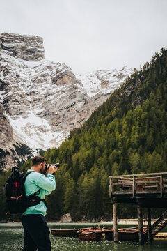 Fot&oacute;grafo con mochila  tomando foto a un lago con estructura de madera, monta&ntilde;a nevadas y bosque. 