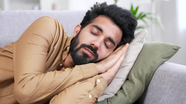 Close up of a young handsome bearded man sleeping during the day while lying on the couch in living room at home. Tired businessman rests dozing with closed eyes on comfortable sofa after work. Siesta