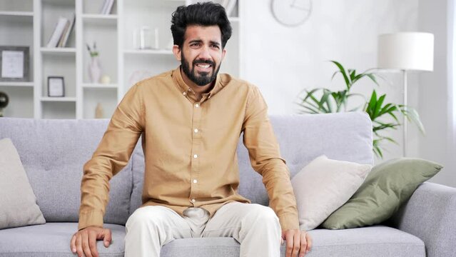 Young bearded male having a panic attack while sitting on sofa in living room at home. A frightened man feels fear, restlessness and anxiety, holds his hands to his chest, breathes hard, is stressed