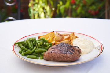 Roasted beef steak from organic agriculture served on the white rustic porcelain plate with boiled green beans and baked potatoes. Lunch served outside in garden restaurant during sunny summer day.