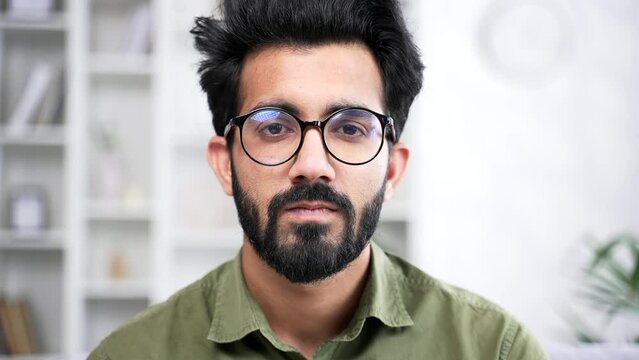 Close up portrait of young handsome bearded businessman in shirt sitting in living room at home office. Head shot of serious coder in glasses. Confident IT programmer developer poses looking at camera