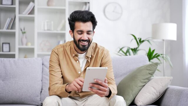 A smiling young freelancer is using, browsing a tablet while sitting on sofa in living room at home office. Bearded entrepreneur is shopping in an online store, chatting or writing message to a client