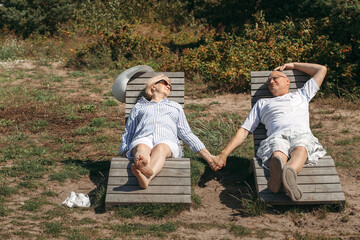 A couple of cute elderly people lie on sunbeds holding hands.