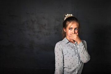 Portrait of scared little kid girl 10 year old in shirt covering mouth posing at black background, looking away. Frightened lovely child lady expresses emotion. Youth gen a concept. Copy ad text space