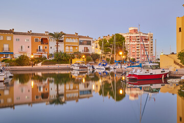 Panoramic view of colorful houses and moored yachts in the evening in Port Saplaya. Light from street lamps, buildings and boats reflects on the smooth surface of the water. Valencia's Little Venice.