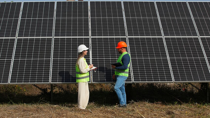 A man and a woman in hard hats and with a laptop are discussing work tasks against the backdrop of solar panels outside. Female environmental engineer talking to investor. Green electricity concept.