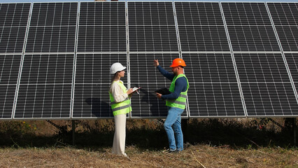 A man and a woman in hard hats and with a laptop are discussing work tasks against the backdrop of solar panels outside. Female environmental engineer talking to investor. Green electricity concept.