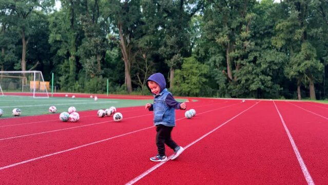 Smiling baby boy in a hood turning around on the red track. Happy boy runs up to many balls scattered on the stadium.