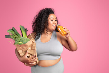 Positive black chubby woman in sportswear holding bag of green vegetables and drinking fresh...