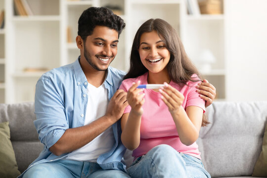 Excited indian couple looking at positive pregnancy test, smiling and enjoying great news, sitting on couch at home
