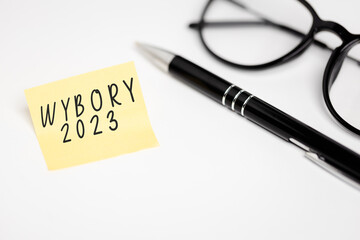 a yellow piece of paper with the inscription "Wybory 2023", next to it a pen and glasses on a white background (selective focus) translation: Election 2023