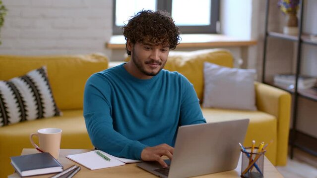 Positive indian guy working distantly at home office interior, typing and editing report on laptop, enjoying remote job