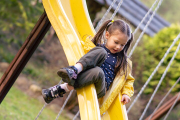 Active girl sitting on slide at playground, looking on way and carefully sliding down, spending leisure on autumn day.