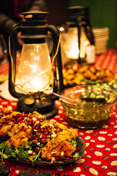 A salad of roast yams with nuts and pomegranate over greens on a red patterned tablecloth next to rustic gas lanterns