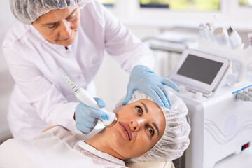 Female cosmetologist doing anti-aging face hardware therapy procedure for young female client in clinic of aesthetic medicine