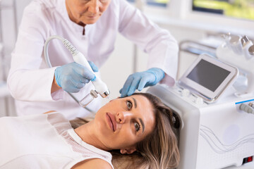Concentrated young female patient undergoing facial procedure with application of atomized nutrients to enhance skin vitality at aesthetic medicine clinic. Modern technologies in hardware cosmetology