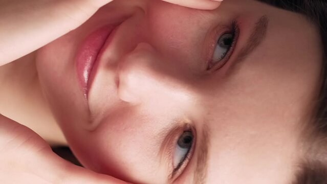 Vertical video. Facial care. Female beauty procedure. Hydration treatment. Pretty smiling woman touching massaging smooth face skin with hands after applying skincare cosmetic.