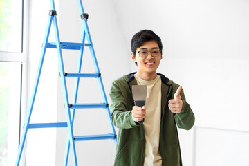 Young Asian man with putty knife showing thumb-up in room