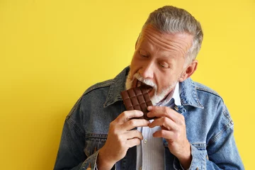Poster Mature man eating tasty chocolate on yellow background, closeup © Pixel-Shot