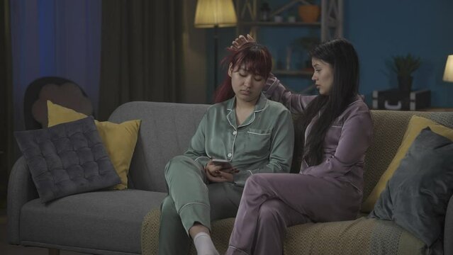 Medium-full video capturing two young women in pajamas sitting on the couch together. One of them is sad about something in her smartphone, her friend, sister thies to cheer her up.
