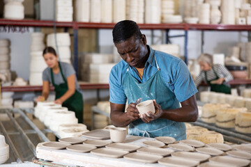 Skillful african-american man ceramist in apron carving new plates in workshop.