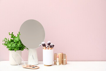 Different makeup products, mirror and houseplant on dressing table near pink wall in room