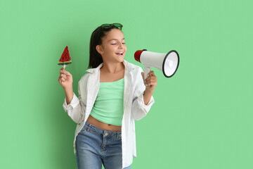 Happy little girl with lollipop in shape of watermelon slice and megaphone on green background