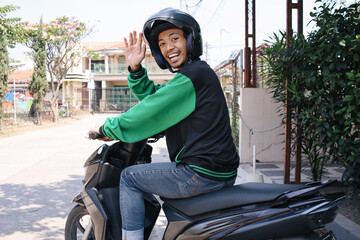 Back view of happy commercial motorcycle taxi driver wearing helmet and uniform jacket waving hand...
