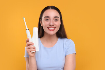 Happy young woman holding electric toothbrush and tube of toothpaste on yellow background