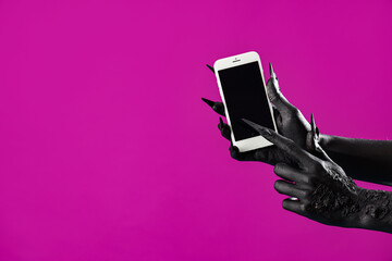 Black hands of witch holding mobile phone with blank screen on purple background. Halloween...