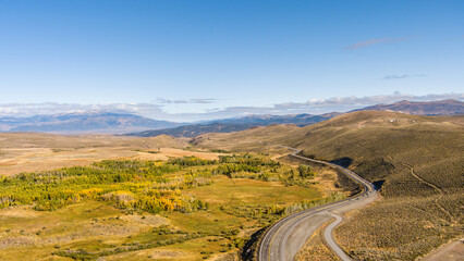 Aerial view of Highway 395 running through Patches of Autumn colored leaves in the California Eastern Sierras landscape with snow capped mountains small clouds and copy space against a bright blue sky