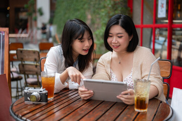 Two cheerful Asian female friends are enjoying talking while watching a video on a tablet together.