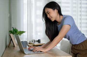 A beautiful Asian woman bending over a table and using her laptop, working in her room.