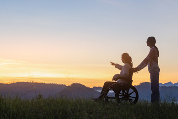 Silhouette woman sitting in wheelchair with daughter looking morning sunrise sky mountain on sunny day - Powered by Adobe