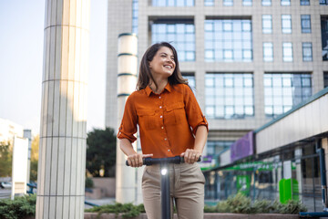 Next generation commute to work using e-scooters by female business persons. Young beautiful woman in a blue jacket smiles and rides an electric scooter to work along office buildings