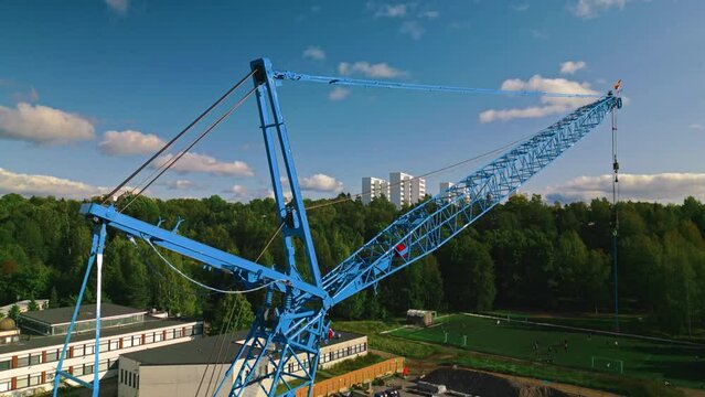Rear strut, front strut, jib and pendant of a blue luffing construction crane against a blue sky and clouds with forest in background. Good view of the main chords. Camera descending.