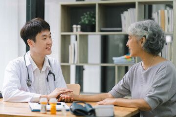 Young Asian male doctor holding hands with a senior patient woman, offering hope and kindness at a modern clinic office.