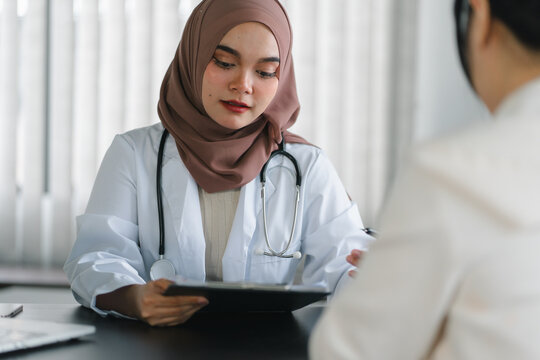Woman patient visiting muslim female doctor at clinic office. Muslim female doctor writes a prescription on a table in a hospital. - Powered by Adobe