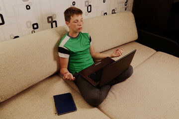 A boy sits on a sofa with a laptop in a yoga pose. The boy practices yoga online. Distance learning yoga. High quality photo