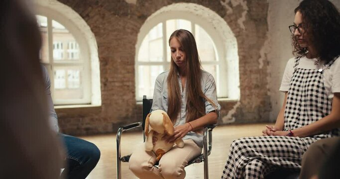 A blonde girl with long hair talks about her problem while holding a soft toy in her hands, and the rest of the group therapy participants support the girl and put their hands on her shoulders