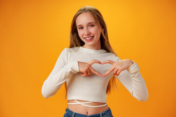 Friendly teen girl shows heart love gesture on yellow background