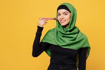 Young smiling arabian asian muslim woman wear green hijab abaya black clothes look camera use brush for make up isolated on plain yellow background. People uae middle eastern islam religious concept.