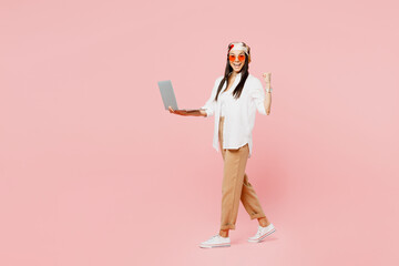 Full body side view young IT latin woman she wearing white shirt casual clothes sunglasses hold use work on laptop pc computer do winner gesture isolated on plain pastel light pink background studio.