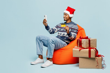 Full body young man in sweater Santa hat posing sit in bag chair use mobile cell phone credit bank...