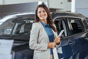 Smiling saleswoman holding tablet PC while looking at camera at new car showroom. Professional car dealer posing proudly at auto showroom, smiling to the camera, holding tablet PC. - obrazy, fototapety, plakaty